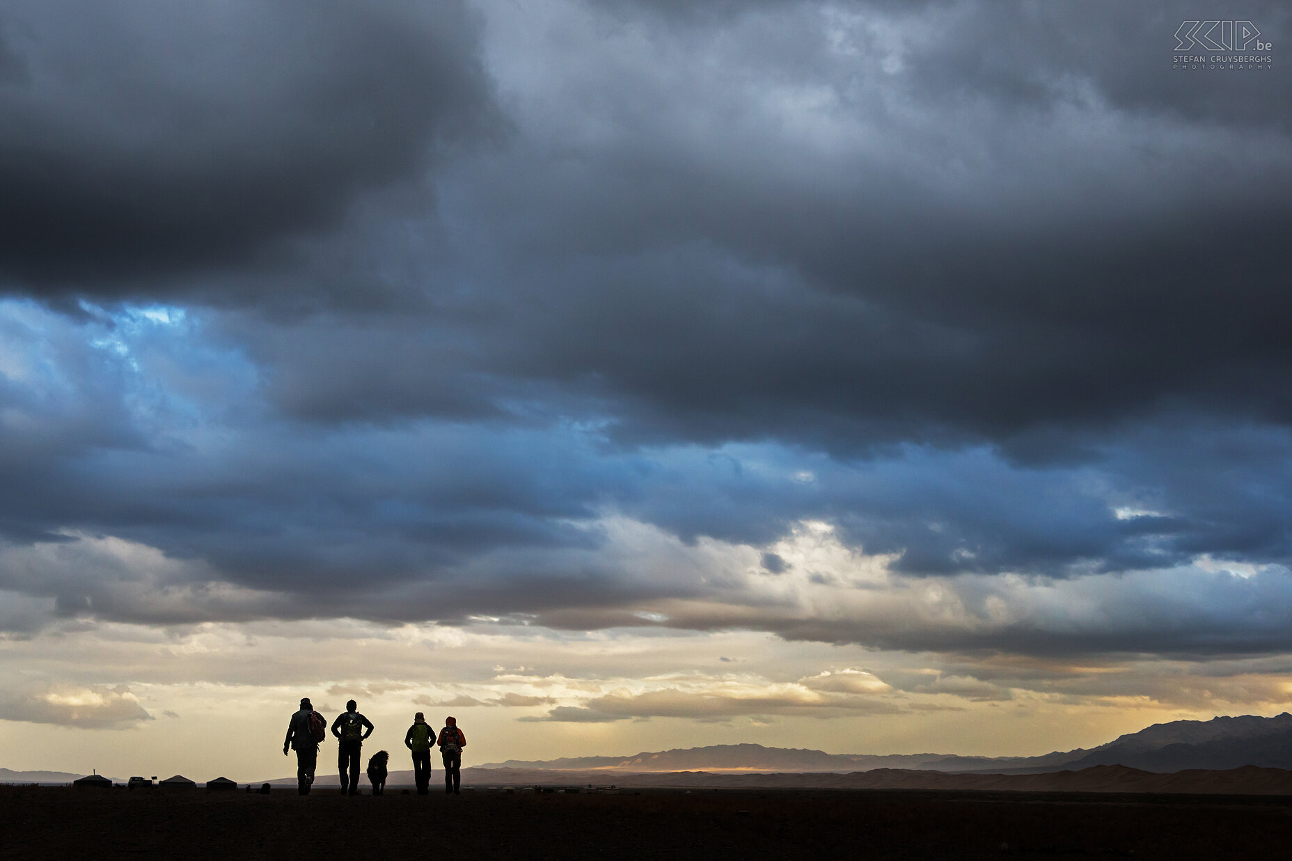 Gobi - Khongoryn Els - Stilte voor de storm We hadden juist een van de hoogste zandduinen van Khongoryn Els in de Gobi woestijn beklommen en we waren op onze terugweg naar de ger van de nomadische familie waar we logeerden. Het werd echter steeds kouder en de lucht werd dreigender. Mijn vrienden en een hond van de nomaden die voorop liepen, leken donkere silhouetten tegen het felle licht in de verte. Dus heb ik nog snel deze foto gemaakt. 10 seconden later kregen we een koude regenbui over ons. Stefan Cruysberghs